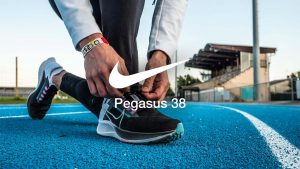Zoom sur les pieds d'un homme faisant ses lacets, avec une paire de Nike Zoom Pegasus 38 au pied. Il est sur une piste d'athlétisme en tartan bleu. Un logo Nike avec l'inscription "Pegasus 38" est au centre de l'image
