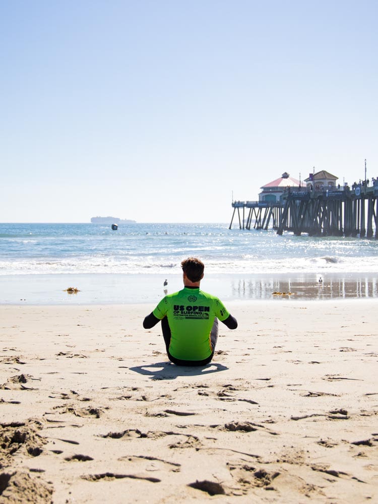 Gaspard Larsonneur de dos, face à l'océan, assis sur la plage de Huntington Beach en Californie, pendant l'US Open of Surfing 2022