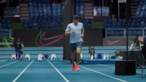 Benjamin Robert en tenue Nike bleue et chaussures rouges, en train de courir sur la piste bleue de l'Arena Stade Couvert de Liévin, la veille de sa course au Meeting de Liévin. Derrière lui, des panneaux publicitaires lumineux affichent des logos Nike.