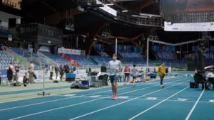 Benjamin Robert en tenue Nike bleue et chaussures rouges, en train de courir sur la piste bleue de l'Arena Stade Couvert de Liévin, la veille de sa course au Meeting de Liévin. Plan large sur lequel on voit les gradins vides.