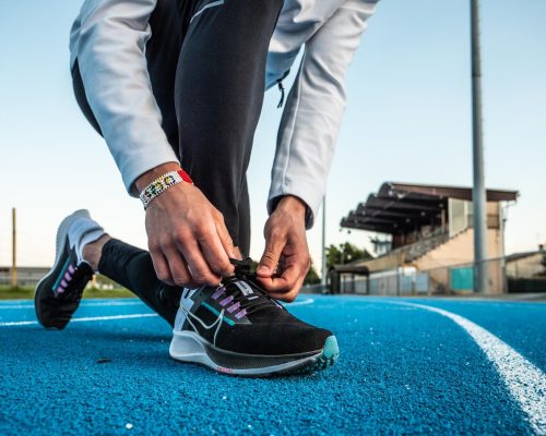 Zoom sur les pieds d'un homme faisant ses lacets, avec une paire de Nike Zoom Pegasus 38 au pied. Il est sur une piste d'athlétisme en tartan bleu. Il y a une tribune de stade en arrière plan.
